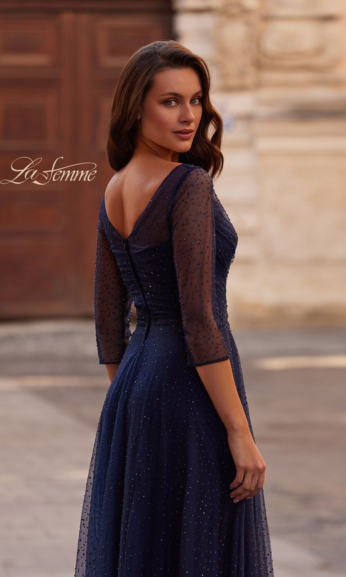 A woman in the Sparkly Sleeved Tulle Formal Dress: La Femme 32460, featuring navy blue tulle, three-quarter sheer dotted sleeves, a ruched A-line silhouette, and a V-back, stands outdoors looking over her shoulder.